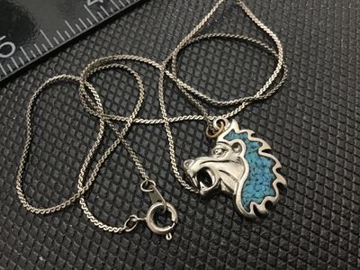 Sale Photo Thumbnail #115: A silver-tone necklace featuring a stylized lion head pendant filled with crushed turquoise chip inlay along the mane. The pendant hangs from a fine woven chain with a spring ring clasp. Shows light vintage wear and age-appropriate patina. Unmarked metal.