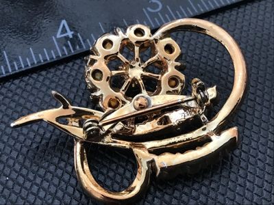 Sale Photo Thumbnail #16: A vintage gold-tone brooch featuring a central floral cluster set with clear round rhinestones surrounded by sweeping curved ribbon-like accents with additional accent stones. Measures approximately 1.75 inches long. Features a rollover safety clasp on th