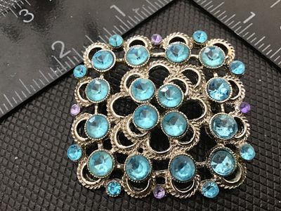 Sale Photo Thumbnail #171: A square vintage silver-tone brooch featuring an openwork filigree-style pattern set with faceted light blue/aqua rhinestones and small purple accent stones. Pin mechanism on the back appears intact and functional. Measures approximately 1.75 inches squar
