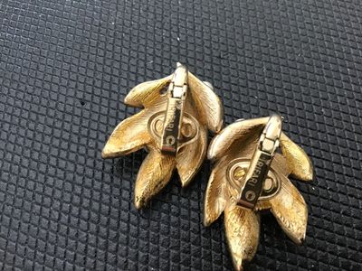 Sale Photo Thumbnail #182: A pair of vintage Crown Trifari clip-on earrings featuring a textured gold-tone leaf design adorned with faux pearl accents. Both earrings are stamped with the Crown Trifari copyright mark on the clip mechanisms. Show light surface wear consistent with ag