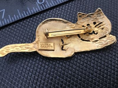 Sale Photo Thumbnail #68: A vintage gold-tone brooch featuring a playful lounging or reclining cat design with openwork facial details and whiskers. Marked 'ULTRA CRAFT ©' on the back. Measures approximately 2.25 inches in length. Standard pin back with rollover safety clasp in g