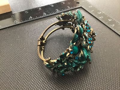 Sale Photo Thumbnail #140: An ornate vintage brass-tone hinged cuff bracelet featuring an elaborate floral design set with sparkling blue rhinestones and green/turquoise-colored cabochons and painted leaf accents. Features a spring hinge closure mechanism. Shows minor finish wear c