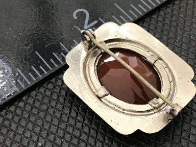 Sale Photo Thumbnail #153: A vintage silver-tone brooch featuring a central oval cabochon stone in a reddish-brown agate or glass tone, surrounded by yellow and brown enamel accents in a Southwestern style frame. Pin back clasp mechanism on the reverse. Shows minor surface wear, en