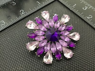 Sale Photo Thumbnail #169: Silver-tone costume brooch styled as a flower or starburst, featuring a dark purple central round rhinestone encircled by light purple and violet teardrop and marquise rhinestone accents. Secured with a standard pin stem and rollover clasp back. Measures 