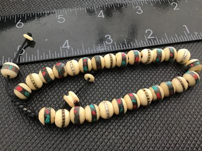 Sale Photo Thumbnail #155: Adjustable woven cord bracelet featuring carved off-white bone beads with inlaid brass bands, red, and turquoise-color stone chips. Includes a sliding knot closure with decorative end beads. Inner circumference is adjustable, beads measure approximately 8