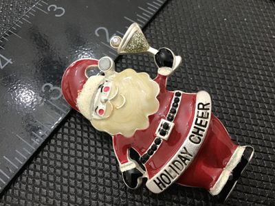 Sale Photo Thumbnail #104: A festive Christmas brooch featuring Santa Claus holding a martini glass with a banner reading 'HOLIDAY CHEER'. Crafted from silver-tone metal with red, white, and cream enameling, accented with black and clear rhinestone details. Measures approximately 2