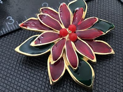 Sale Photo Thumbnail #26: Gold-tone metal brooch shaped like a Christmas poinsettia flower, featuring deep red and green enamel fill with three red faux-pearl/bead accents at the center. Measures approximately 2.5 inches in diameter. Has a textured back with a rollover safety clas