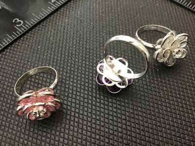 Sale Photo Thumbnail #126: A set of three silver-tone costume jewelry rings featuring layered flower designs with rhinestone and colored crystal details in pink, purple, and clear. Rings are lightweight metal with thin bands, measuring approximately size 4 to 6. Good condition with