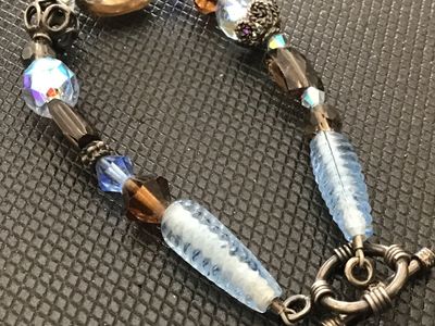 Sale Photo Thumbnail #162: Artisan beaded bracelet featuring faceted crystal beads, smoky brown and iridescent light blue glass accents, filigree metal beads, and a ornate toggle clasp. Measures approximately 7.5 to 8 inches in length. In good pre-owned condition with no visible da