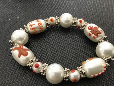 Sale Photo Thumbnail #159: A festive holiday stretch bracelet featuring faux pearl beads accented with painted gingerbread man motifs, a gingerbread house, and text details ("Muffin Man", "Knows"). Separated by silvertone textured spacer beads. Appears to be in good pre-owned condi