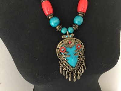 Sale Photo Thumbnail #190: Boho style statement necklace featuring a large ornate brass-tone pendant with faux turquoise, coral-colored flower details, and dangling tassels. Strung with turquoise-blue and bright coral chunky beads alongside dark wood beads near the back. Features a
