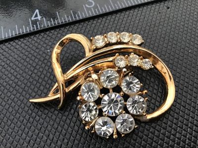 Sale Photo Thumbnail #15: A vintage gold-tone brooch featuring a central floral cluster set with clear round rhinestones surrounded by sweeping curved ribbon-like accents with additional accent stones. Measures approximately 1.75 inches long. Features a rollover safety clasp on th