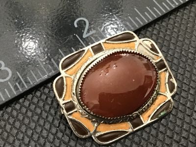 Sale Photo Thumbnail #152: A vintage silver-tone brooch featuring a central oval cabochon stone in a reddish-brown agate or glass tone, surrounded by yellow and brown enamel accents in a Southwestern style frame. Pin back clasp mechanism on the reverse. Shows minor surface wear, en