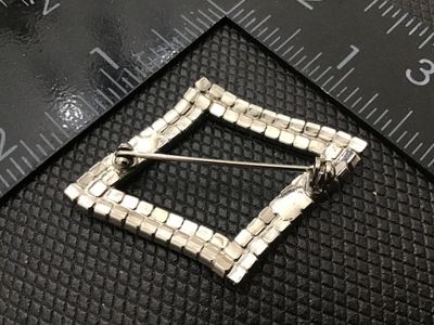 Sale Photo Thumbnail #103: A vintage diamond-shaped brooch featuring two rows of prong-set clear sparkling rhinestones set in silver-tone metal. Measures approximately 1.5 inches long by 1.25 inches wide. Features a standard pin back with rotating C-clasp mechanism. Good vintage co