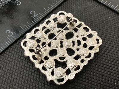 Sale Photo Thumbnail #172: A square vintage silver-tone brooch featuring an openwork filigree-style pattern set with faceted light blue/aqua rhinestones and small purple accent stones. Pin mechanism on the back appears intact and functional. Measures approximately 1.75 inches squar