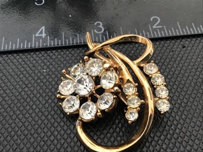 Sale Photo Thumbnail #17: A vintage gold-tone brooch featuring a central floral cluster set with clear round rhinestones surrounded by sweeping curved ribbon-like accents with additional accent stones. Measures approximately 1.75 inches long. Features a rollover safety clasp on th