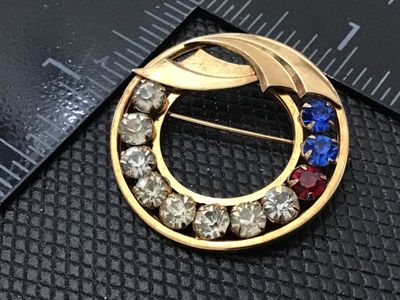 Sale Photo Thumbnail #135: A vintage circular openwork brooch featuring clear, blue, and red prong-set rhinestones along the lower rim, topped with a textured gold-tone leaf or ribbon accent. Marked 1/20 12K GF on the reverse. Pin mechanism is intact. Measures approximately 1 inch 