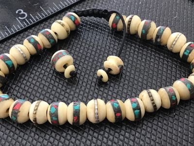 Sale Photo Thumbnail #154: Adjustable woven cord bracelet featuring carved off-white bone beads with inlaid brass bands, red, and turquoise-color stone chips. Includes a sliding knot closure with decorative end beads. Inner circumference is adjustable, beads measure approximately 8