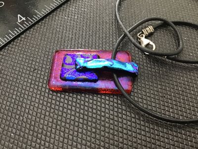 Sale Photo Thumbnail #110: Artisan dichroic fused glass pendant featuring layered iridescent purple, blue, and magenta tones. The rectangular pendant measures approximately 1.5 inches long and hangs from a black cord necklace with a metallic lobster claw clasp. In good pre-owned co