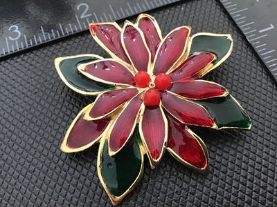 Sale Photo Thumbnail #24: Gold-tone metal brooch shaped like a Christmas poinsettia flower, featuring deep red and green enamel fill with three red faux-pearl/bead accents at the center. Measures approximately 2.5 inches in diameter. Has a textured back with a rollover safety clas
