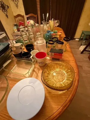 Sale Photo Thumbnail #12: Large assortment of kitchen glassware and tableware, including clear Pyrex baking dishes, colorful coffee mugs, pressed amber glass bowls, and cobalt blue-rimmed drinking glasses.