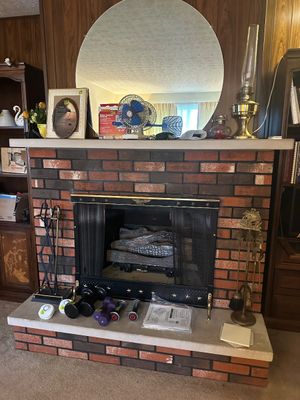Sale Photo Thumbnail #39: Traditional brick fireplace with a mantle featuring a large circular mirror, small electric fan, oil lamps, and miscellaneous decor, alongside hand weights set on the hearth.