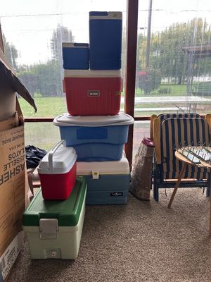 Sale Photo Thumbnail #20: Collection of vintage and modern plastic beverage coolers and chests stacked in a sunroom, featuring brands like Thermos and Gott in various colors and sizes.