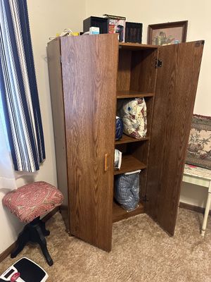 Sale Photo Thumbnail #3: Tall wood-grain laminate storage cabinet with double doors and interior shelves containing linens, alongside an upholstered vanity stool.