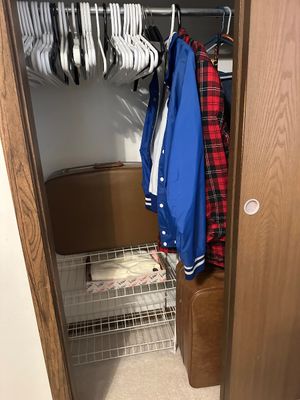 Sale Photo Thumbnail #7: A bedroom closet containing two vintage brown suitcases, a blue windbreaker, a red plaid jacket, a wire shelving rack, and various hangers.