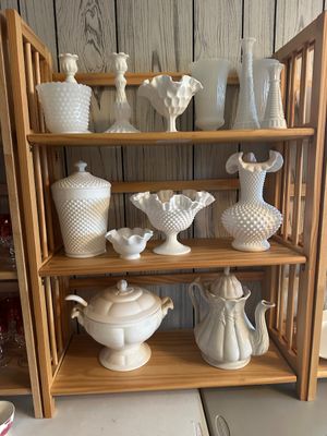 Sale Photo Thumbnail #38: Collection of vintage milk glass tableware and decorative items displayed on a light wood shelving unit. Includes Fenton hobnail vases, ruffled compotes, candlesticks, a covered jar, and cream-colored ceramic serving pieces.
