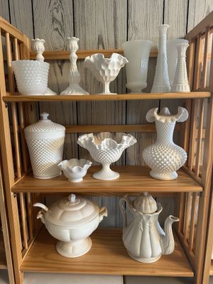 Sale Photo Thumbnail #60: Collection of vintage white milk glass items displayed on a light wood shelf, including Fenton hobnail vases, candy dishes, candlesticks, and covered serveware.