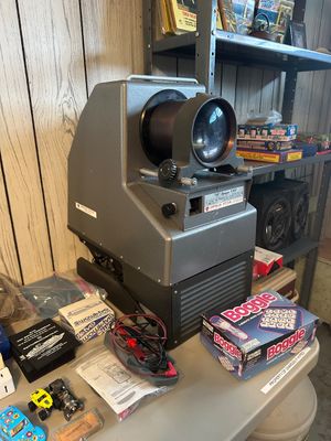 Sale Photo Thumbnail #48: Vintage American Optical Company AO Opaque 1000 projector, sitting alongside a vintage Boggle board game and various small collectibles on a table.