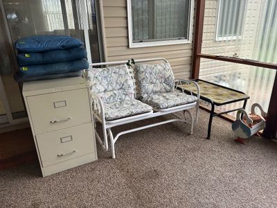 Sale Photo Thumbnail #19: White metal patio glider bench with floral cushions, shown alongside a beige two-drawer file cabinet, a sunflower-accented coffee table, and a decorative wooden goose planter.