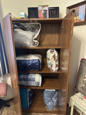 Sale Photo Thumbnail #4: Tall wood veneer armoire cabinet filled with bagged quilts and bedding, topped with compact space heaters.