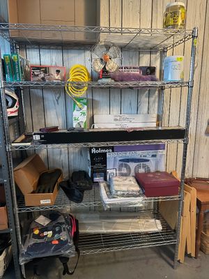 Sale Photo Thumbnail #51: Five-tier metal wire shelving unit organized with assorted household goods, including a vintage desk fan, boxed Holmes window fan, CD-R spindle, auto accessories, and a space-themed backpack.