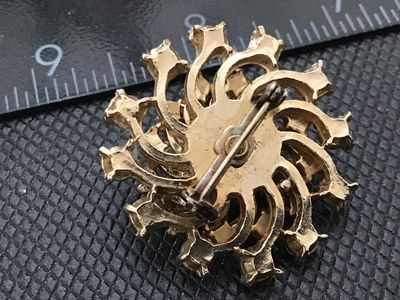 Sale Photo Thumbnail #157: A vintage gold-tone starburst brooch featuring a bright green center rhinestone surrounded by clear rhinestones, faux pearls, and purple rhinestone accents along the outer spiral design. Measures approximately 1.25 inches in diameter. Shows light vintage 