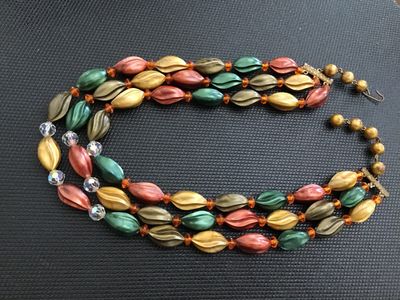 Sale Photo Thumbnail #189: A vintage 3-strand beaded necklace featuring swirl/leaf-molded plastic beads in earth tones (green, gold, bronze, terracotta/coral), accented with faceted crystal and amber spacer beads. Includes decorative metal bar connectors and a hook-and-tail clasp e