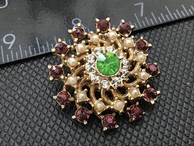 Sale Photo Thumbnail #156: A vintage gold-tone starburst brooch featuring a bright green center rhinestone surrounded by clear rhinestones, faux pearls, and purple rhinestone accents along the outer spiral design. Measures approximately 1.25 inches in diameter. Shows light vintage 
