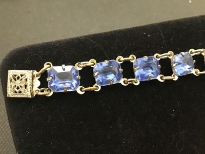 Sale Photo Thumbnail #205: Vintage costume jewelry link bracelet featuring emerald-cut light blue glass stones set in open-backed claw prongs. Includes a decorative filigree push-in box clasp with safety tongue. Measures approximately 7.25 inches in overall length. Metal shows surf