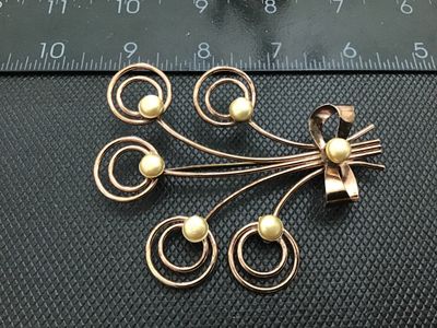 Sale Photo Thumbnail #120: A vintage wirework brooch designed as a floral spray or bouquet, tied with a ribbon bow. Crafted in a rose gold tone metal with swirling wire details topped with Cream-colored faux pearl accents. Measures approximately 2.5 inches wide. Shows light age-app