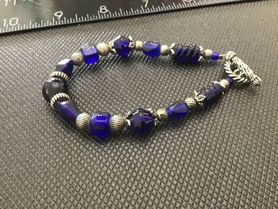 Sale Photo Thumbnail #146: Beaded bracelet featuring vibrant cobalt blue art glass beads in various shapes and textures, interspersed with ornate silver-tone spacer beads, bead caps, and a decorative rope-style toggle clasp. Measures approximately 7 to 7.5 inches long. In good cond