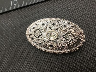 Sale Photo Thumbnail #147: Oval vintage silver-tone brooch featuring an openwork filigree design set with small pavé rhinestones and a central oval crystal. Measures approximately 2 inches wide based on the ruler in photos. Back features a standard pin stem with a rolling c-clasp 
