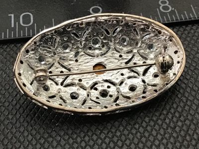 Sale Photo Thumbnail #148: Oval vintage silver-tone brooch featuring an openwork filigree design set with small pavé rhinestones and a central oval crystal. Measures approximately 2 inches wide based on the ruler in photos. Back features a standard pin stem with a rolling c-clasp 