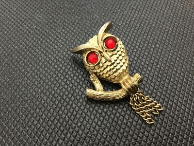 Sale Photo Thumbnail #194: A vintage gold-tone owl brooch by Avon featuring red rhinestone eyes and dangling chain accents from the branch perch. Marked 'Avon ©' on the back. Includes a standard rollover safety clasp pin back. In good vintage condition with minor surface wear cons