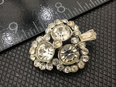 Sale Photo Thumbnail #150: Small vintage silver-tone rhinestone brooch shaped like a shamrock or clover with a small stem. Set with three larger round chaton rhinestones, surrounded by halo accents and a baguette stone on the stem. Back features a pin mechanism. Shows light surface