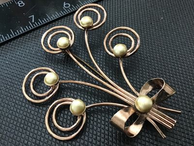 Sale Photo Thumbnail #119: A vintage wirework brooch designed as a floral spray or bouquet, tied with a ribbon bow. Crafted in a rose gold tone metal with swirling wire details topped with Cream-colored faux pearl accents. Measures approximately 2.5 inches wide. Shows light age-app