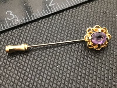 Sale Photo Thumbnail #104: A vintage gold-tone stick pin featuring an openwork filigree setting with an oval faceted purple amethyst-colored stone. Comes complete with a matching gold-tone protective end cap. Total length is approximately 2.5 to 3 inches long based on ruler marking