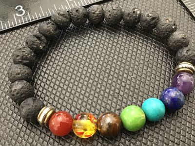Sale Photo Thumbnail #100: Stretch beaded bracelet featuring black porous lava rock/lava stone beads and seven multi-colored gemstone-style chakra accent beads separated by small metal spacer beads. Appears to be in good pre-owned condition with light wear.