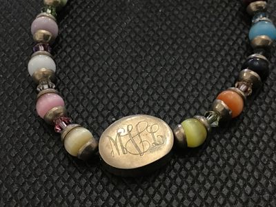 Sale Photo Thumbnail #213: A beaded toggle-clasp bracelet featuring colorful cat's eye synthetic or glass beads alternating with faceted crystal spacers and silver-tone accents. Includes an oval central charm with an engraved script monogram ("MJL"). The toggle ring is stamped "925