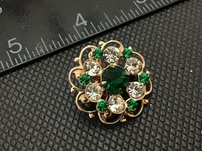Sale Photo Thumbnail #214: A vintage gold-tone openwork filigree style brooch set with green and clear rhinestones. Features a central large faceted green rhinestone surrounded by clear and smaller green accent stones. Pin back mechanism on reverse. Measures approximately 1 inch in